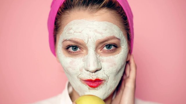 Close up of a girl with a face mask and a towel on her head that eats an apple on a pink background. Beautiful girl with cosmetic mask on her face. Woman wearing face mask. Face care concept.
