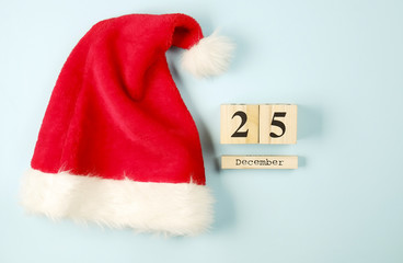 Obraz na płótnie Canvas Colorful Santa Claus hat with fluffy pum-pum laying on blue background. Wooden cube calendar showing Christmas date 25 December. Close up, copy space, top view, flat lay. Holidays preparation concept.