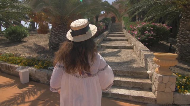 Beautiful woman running up the stairs near luxury villa in Italy. Nice hotel territory. Girl walking and dressed in elegant white dress and hat. Slow motion. Good weather, blue sky. Following camera