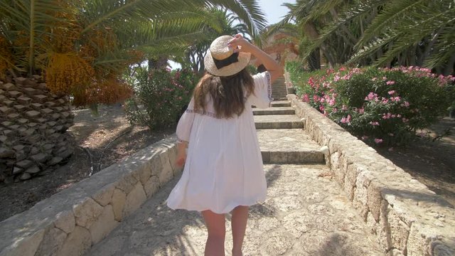 Beautiful woman running up the stairs near luxury villa in Italy. Nice hotel territory. Girl walking and dressed in elegant white dress and hat. Slow motion. Good weather, blue sky.