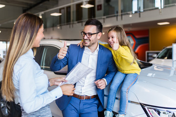 Naklejka premium Handsome young man with his cute little daughter choosing a new car at car showroom.