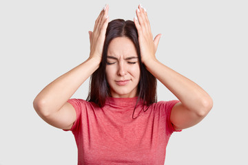 Photo of brunette woman with desperate look, keeps both hands on head, suffers from terrible headache, dressed in casual t shirt, poses against white background. People and tiredness conncept