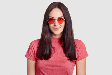 Indoor shot of stylish brunette girl wears trendy red round sunglasses, casual t shirt, ready to have walk during sunny day, isolated over white background. People, fashion and lifestyle concept
