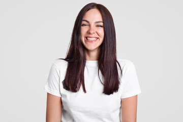 Obraz premium Cheerful satisfied brunette lady has toothy smile, being in good mood, dressed in casual t shirt, expresses positive emotions, isolated over white background. People and facial expressions concept