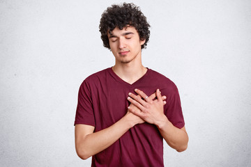 Thankful young boy keeps hands on heart, being touched by heart piercing story, keeps eyes closed,...