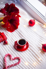 Red cup with espresso coffee on white knitted plaid surrounded with winter decor and garland lights.