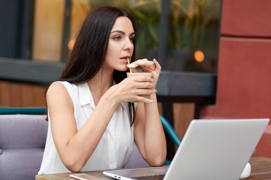 Thoughtful businesswoman has coffee break in outdoor cafeteria, uses laptop computer for distant work, waits for call on smart phone, has concentrated loook into distance, thinks about something