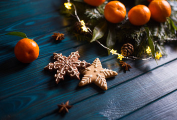 Two Christmas cookies in form of star and snowflake with mandarins, spruce branches, garland lights and star anise on navy rustic wooden table.