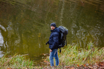 a tourist in a jacket and jeans looks at the camera near the river on the background of an autumn landscape