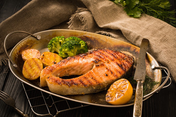 Grilled salmon steak with potatoes, broccoli, lemon and herbs in old roasted metal plate on a wooden table rustic stile