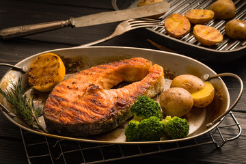 Grilled salmon steak with potatoes, broccoli, lemon and herbs in old roasted metal plate on a...