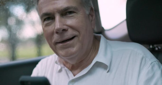 A mature Caucasian man using Uber or Lyft ridesharing checks the mobile app as he gets in the car and engages the driver in friendly conversation.