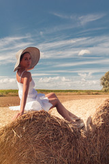 Naklejka na ściany i meble The girl in a hat sits on a haystack in the field.