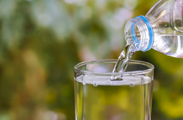 Pouring of clear drink water from bottle into the glass on blurred green nature background with copy space for text.