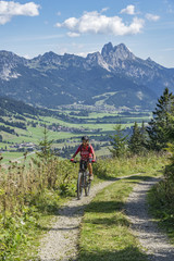 Fototapeta na wymiar nice and active senior woman, riding her e-mountainbike in the Tannheim valley, Tirol, Austria with the village of Tannheim and famous summits Gimpel and Rote Flueh