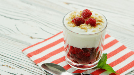 Glass filled with fresh red raspberry and yogurt topped with crispy granola and berries served on napkin with spoon