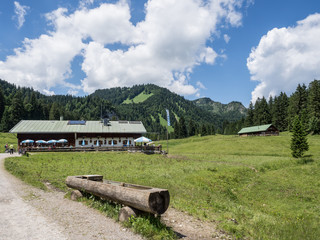 Almwiese mit Wanderweg und Kuhtränke unter weiss blauem Himmel, Bayern