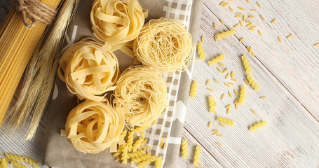 From above view of different type of uncooked pasta laid on tablecloth on white wooden background 