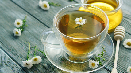 Closeup shot of glass mug on saucer filled with herbal tea and tiny camomile flowers on wooden table with jar of honey