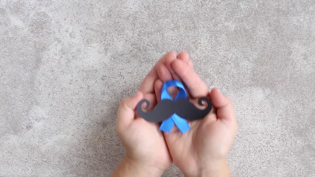 Mustache pattern with blue ribbon symbol in child's hands. Movember concept. Prostate Cancer and men's health awareness.