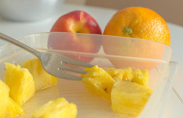Pineapple slices in a plastic can, picked up with a metal fork. An orange and a nectarine fruit in the background, lying on a white table. Healthy fruits as a quick meal in between.