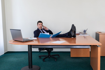 Portrait of young businessman sitting with his feet on the desk in the office talking on the mobile phone while verifying data from a file