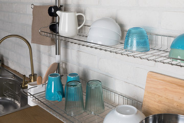 Clean dishes drying on metal dish rack on light background. Kitchen utensils and dishware on wooden shelf. Kitchen interior background.Text space.