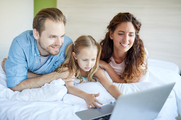 Young family of three lying on bed with lapto pin front and watching movie on weekend