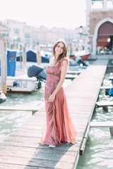 Beautiful young girl in a pink dress standing on a wooden bridge between the Venetian canal and enjoying the city.