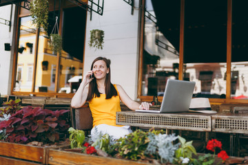 Happy girl in outdoors street cafe sitting at table with laptop pc computer, talking on mobile...