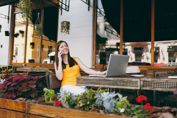 Happy girl in outdoors street cafe sitting at table with laptop pc computer, talking on mobile phone, conducting pleasant conversation, in restaurant during free time. Mobile office freelance concept