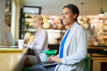 Young busy female in smart casual typing on laptop keypad while sitting by table and getting inspired