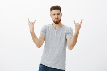 Fototapeta premium Let us rock this party. Portrait of enthusiastic handsome and stylish male model with beard and moustache showing rock-n-roll gesture pouting and frowning enjoying concert and music against white wall