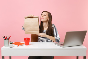 Woman hold brown clear empty blank craft paper bag, sniffing smell work at office with pc laptop isolated on pink background. Food products delivery courier service from shop or restaurant to office.