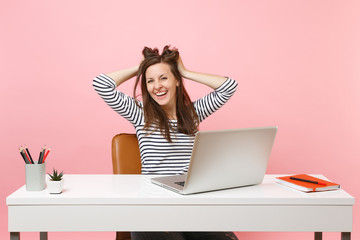 Young laughing woman clinging to hair, head finish working complete project with pc laptop while sitting at office isolated on pastel pink background. Achievement business career concept. Copy space.