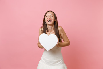 Fototapeta na wymiar Portrait of laughing happy bride woman in beautiful white wedding dress standing holding white heart with copy space isolated on pastel pink background. Wedding celebration concept. Advertising area.