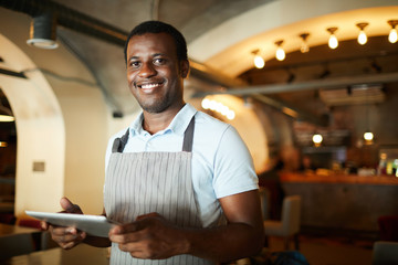 Young waiter in apron holding touchpad while making changes in menu of restaurant
