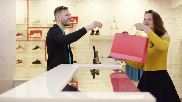 Cheerful customer is using a credit card to pay for purchased clothes while standing at cashier's desk. Shop assistant is accepting transaction and giving paper bags.
