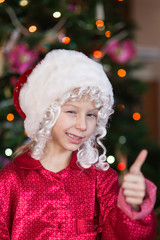 Santa claus  show Ok with a finger near to christmas tree. Small girl in hat of Santa make funny...