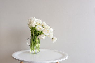 White double tulips in glass jug on small round table against neutral wall background (selective focus)
