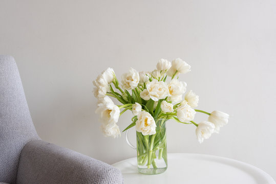 Fototapeta Close up of white double tulips in glass jar on small white table next to retro grey chair (selective focus)
