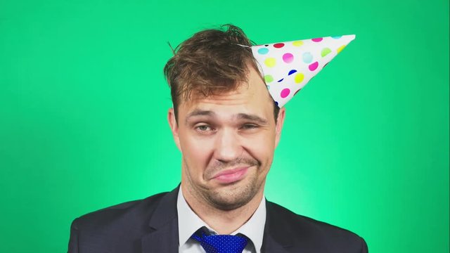 sleepy, disheveled young man in a suit and tie, with a hangover, in a festive cap, yawns and looks at the camera. 4k, green background, slow motion. emotions