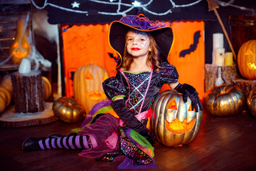 Happy Halloween. A little beautiful girl in a witch costume celebrates at home in an interior with pumpkins and cardboard magic house on the background