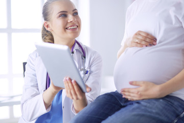 Smiling woman doctor shows pictures on the tablet to pregnant young woman at hospital.