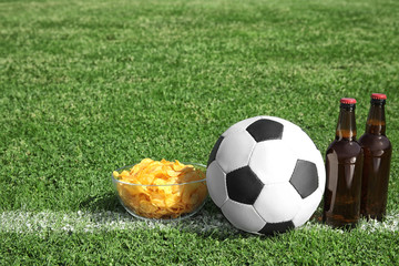 Soccer ball with beverage and chips on green football field grass. Space for text