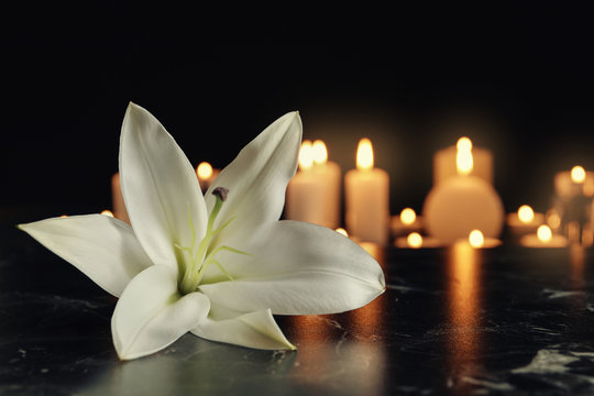 White lily and blurred burning candles on table in darkness, space for text. Funeral symbol