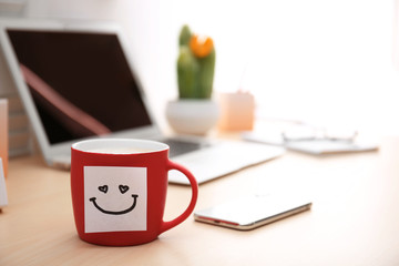 Sticky note with funny face attached to cup of coffee on office table. Space for text