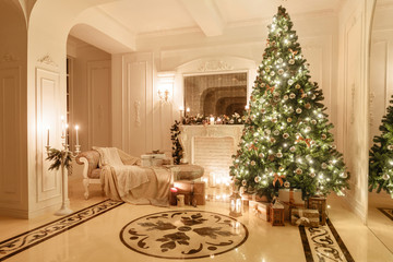 classical interior of a white room. Christmas evening by candlelight. classic apartments with a white fireplace, decorated christmas tree, sofa, large windows and chandelier.