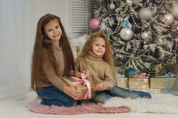 Naklejka premium Two little sisters are waiting for Christmas. Girls are sitting at Christmas tree, smiling and holding gifts. They are dressed in jeans and sweaters. Happy childhood.