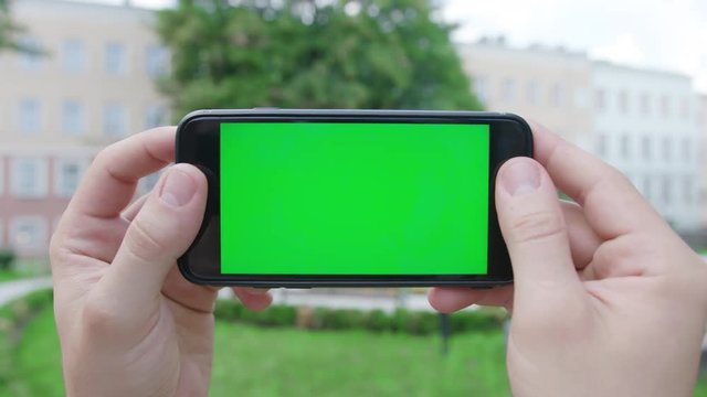Two hands holding a phone with a green screen in a horizontal position in a public park. Close-up shot. Soft focus
